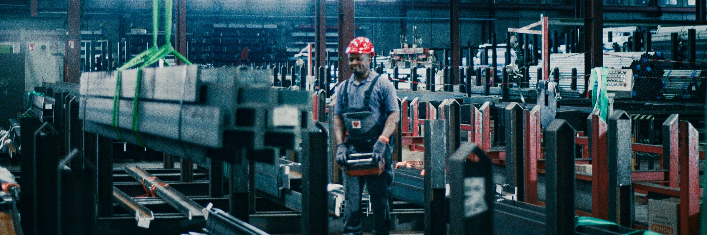 A worker in a hard hat and gloves carries a steel part through the warehouse, between racks of steel sections.