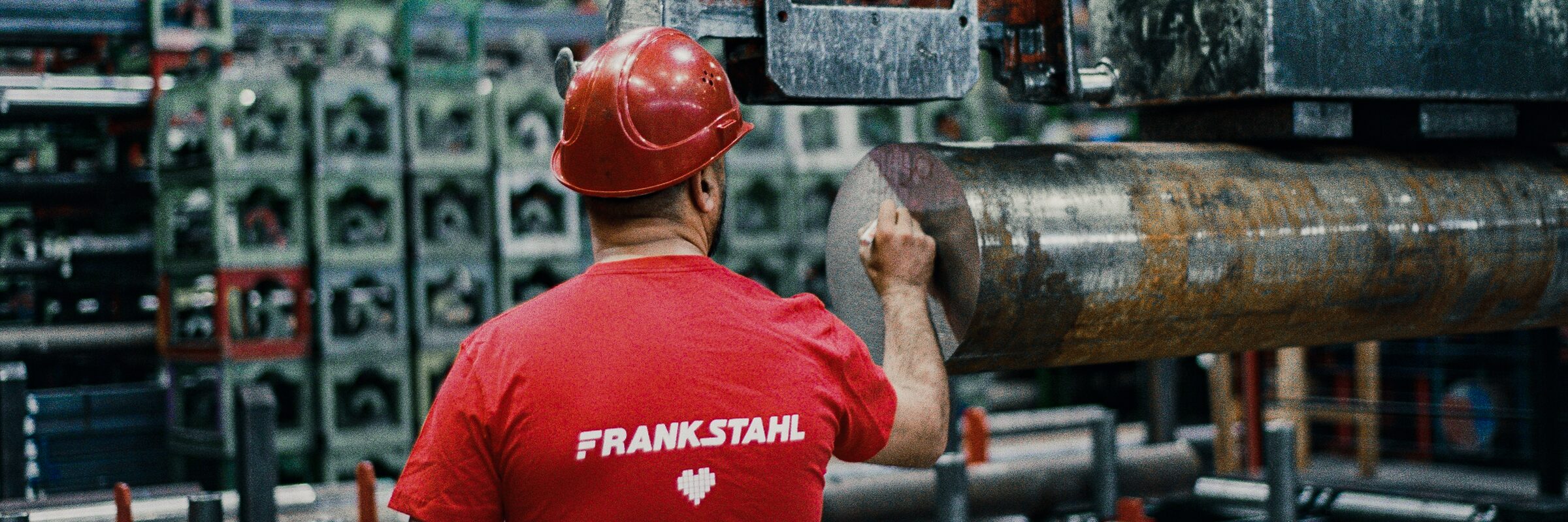 Seen from behind, a worker in a hard hat and FRANKSTAHL shirt guides a round steel bar under the lifting magnet.